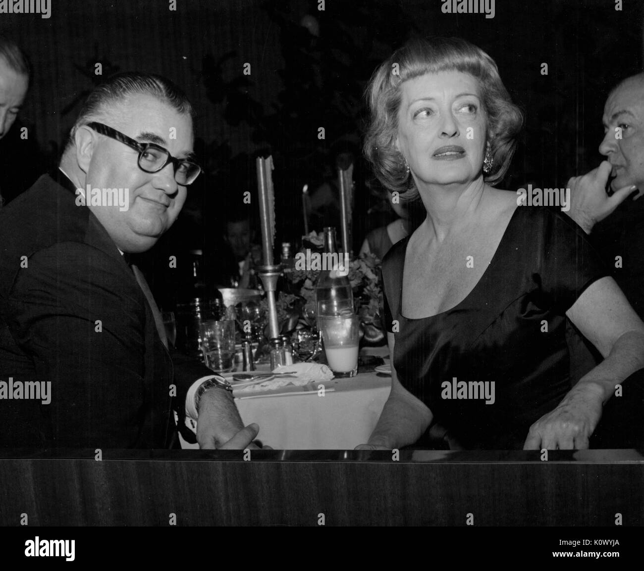 Bette Davis, sitzen bei einem Bankett Tisch mit einem nicht identifizierten Mann, um in ihren Stuhl und schaut, um ihr Recht, in einem dunklen Zimmer bei einem Abendessen, Hollywood, Kalifornien, Hollywood, Kalifornien, 1950. Photo credit Smith Collection/Gado/Getty Images. Stockfoto