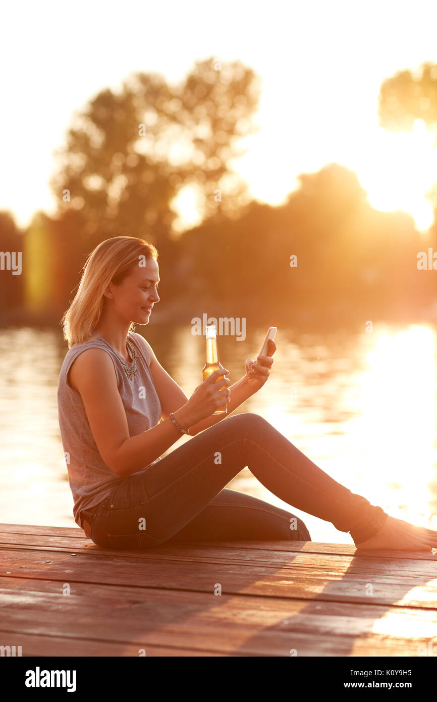Frau auf der Suche nach Handys auf dem Dock und erfrischt mit Drink bei Sonnenuntergang Stockfoto