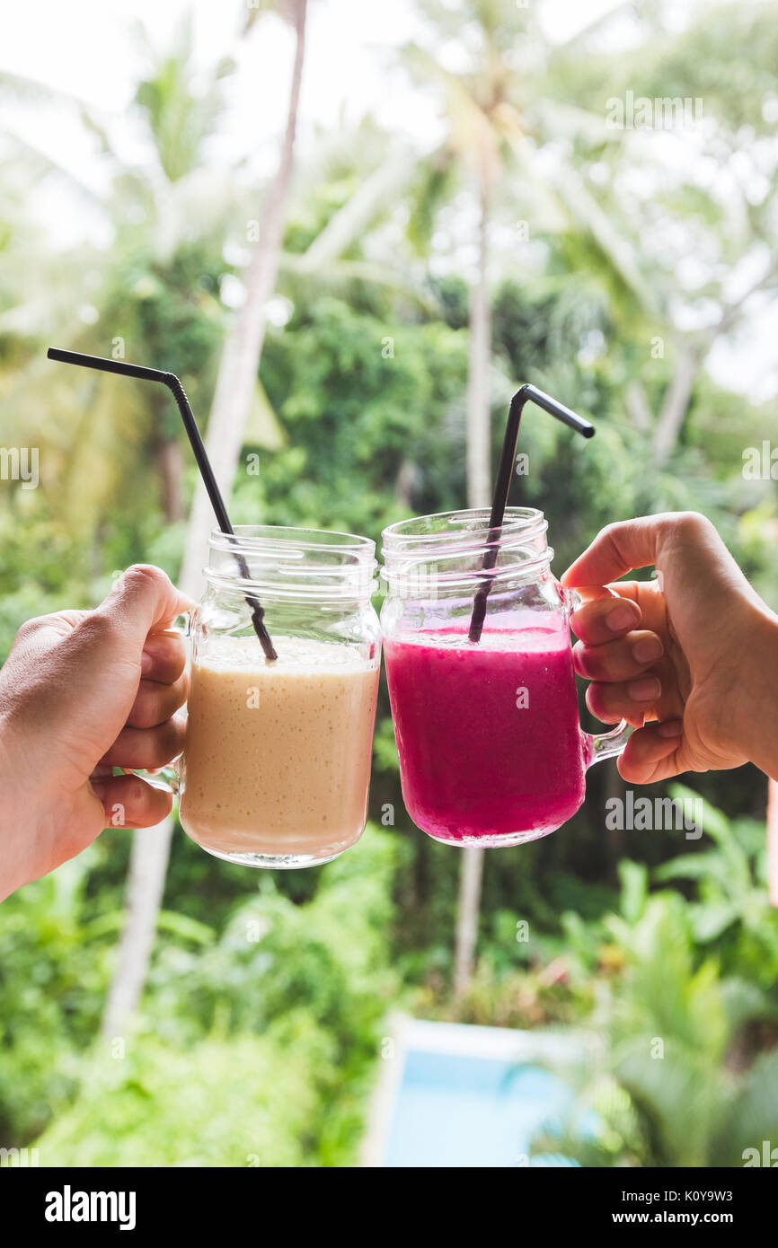 Zwei bunte Obst schüttelt in Händen. Sommer und tropische Stimmung. Kalte Getränke gemischt, Banane und Dragon Fruit Smoothie. Clink Gläser durch Paar Hände Stockfoto