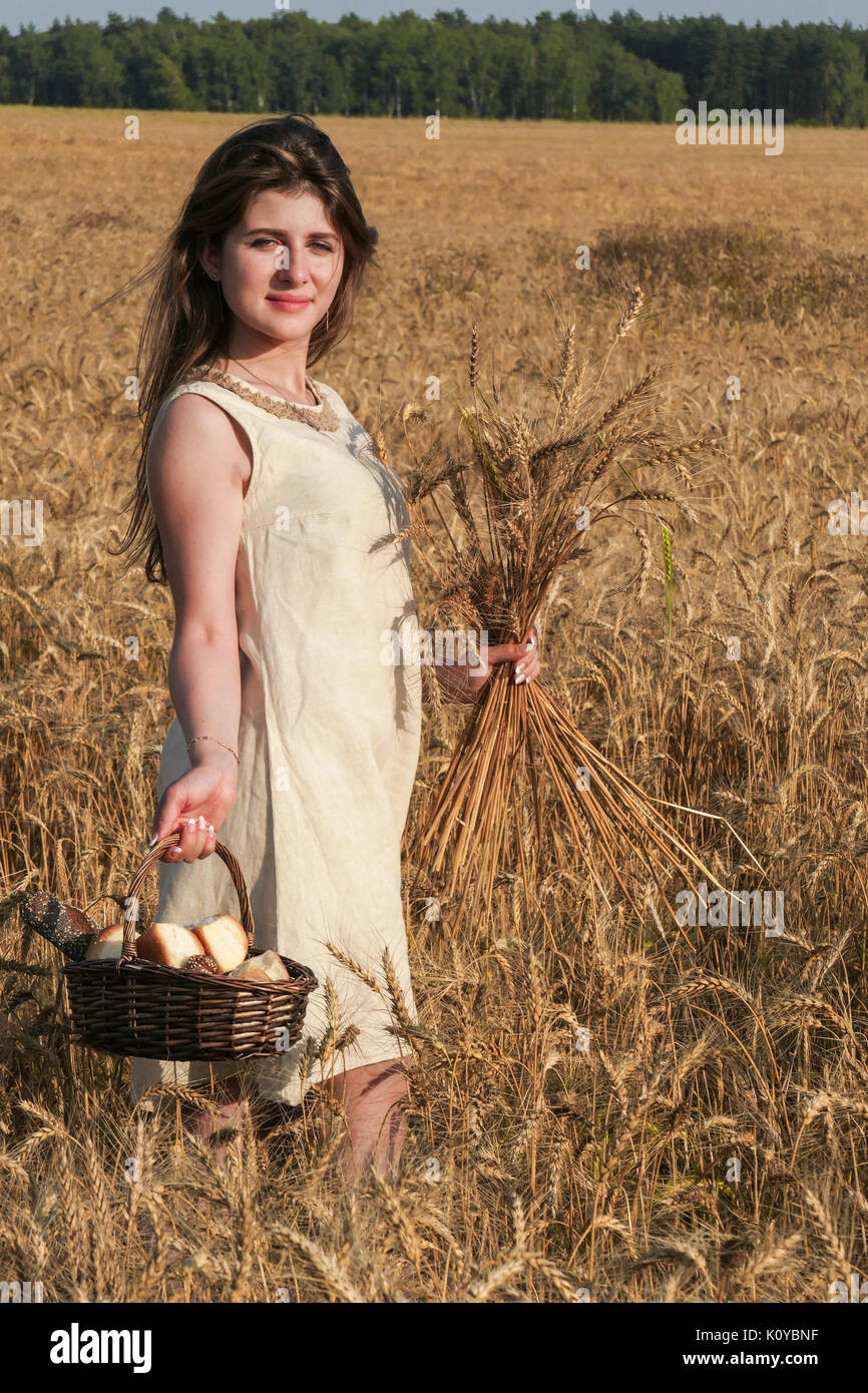 Junge attraktive Frau in wunderschönen natürlichen Kleid wandern mit Korb mit Brot in der goldene Weizen Feld während der Sonnenschein. Stockfoto