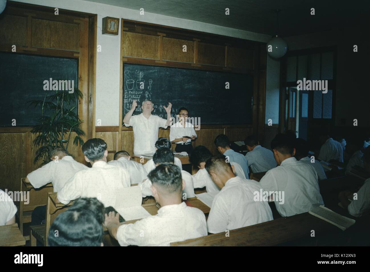 Kaukasische Prediger mit seinen Armen in der Luft, vor einer Tafel an der Vorderseite eines Klassenzimmers in einer missionarischen Kirche, Schreiben auf der Tafel lesen Reinheit und Vergebung, mit einem Bild einer Blume, eine große Gruppe von japanischen Männern tragen weiße Kleidung sitzen im Klassenzimmer und Notizen, Japan, 1952. Stockfoto