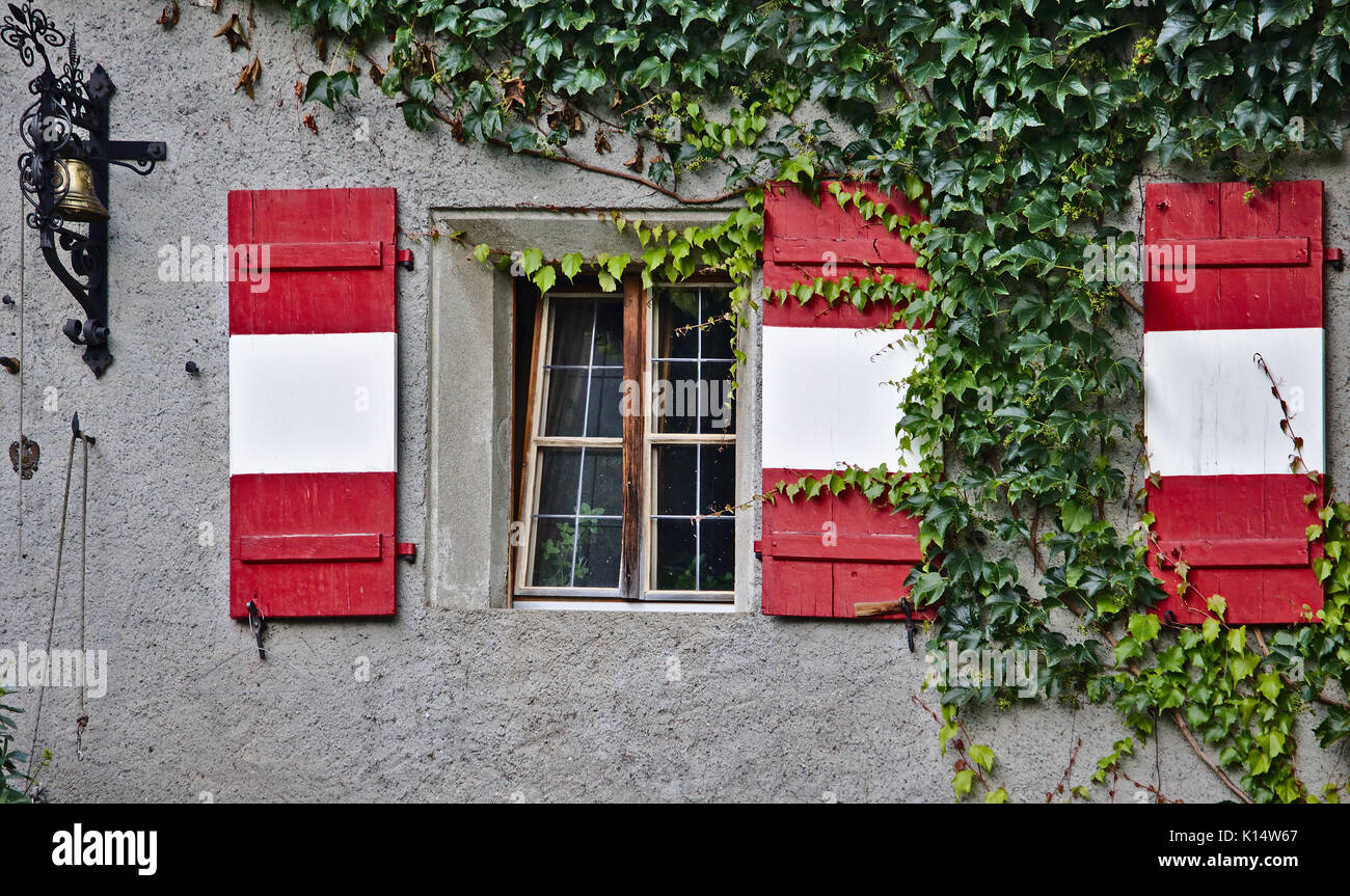 Verputzte Wand mit Fenster, grünen Reben, antike Bell und roten und weißen Fensterläden aus Holz Stockfoto