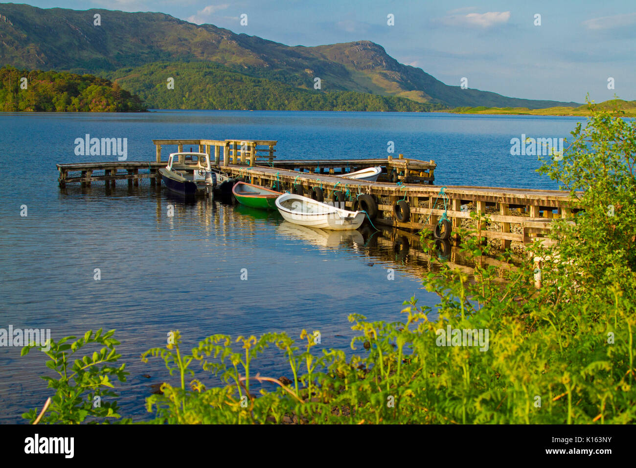 Ruhige, blaue Wasser des Loch Morar gesäumt mit zerklüfteten Bergen und mit Booten an der alten Holzsteg unter blauem Himmel Schottland günstig Stockfoto