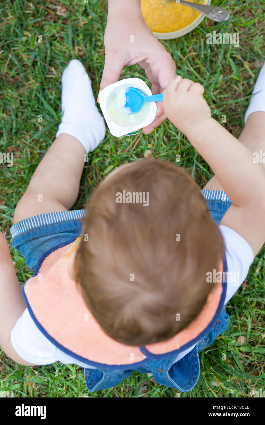 2-jähriger Junge einen Joghurt essen, sitzen auf dem Gras. Er wird von seiner Mutter geholfen worden Stockfoto