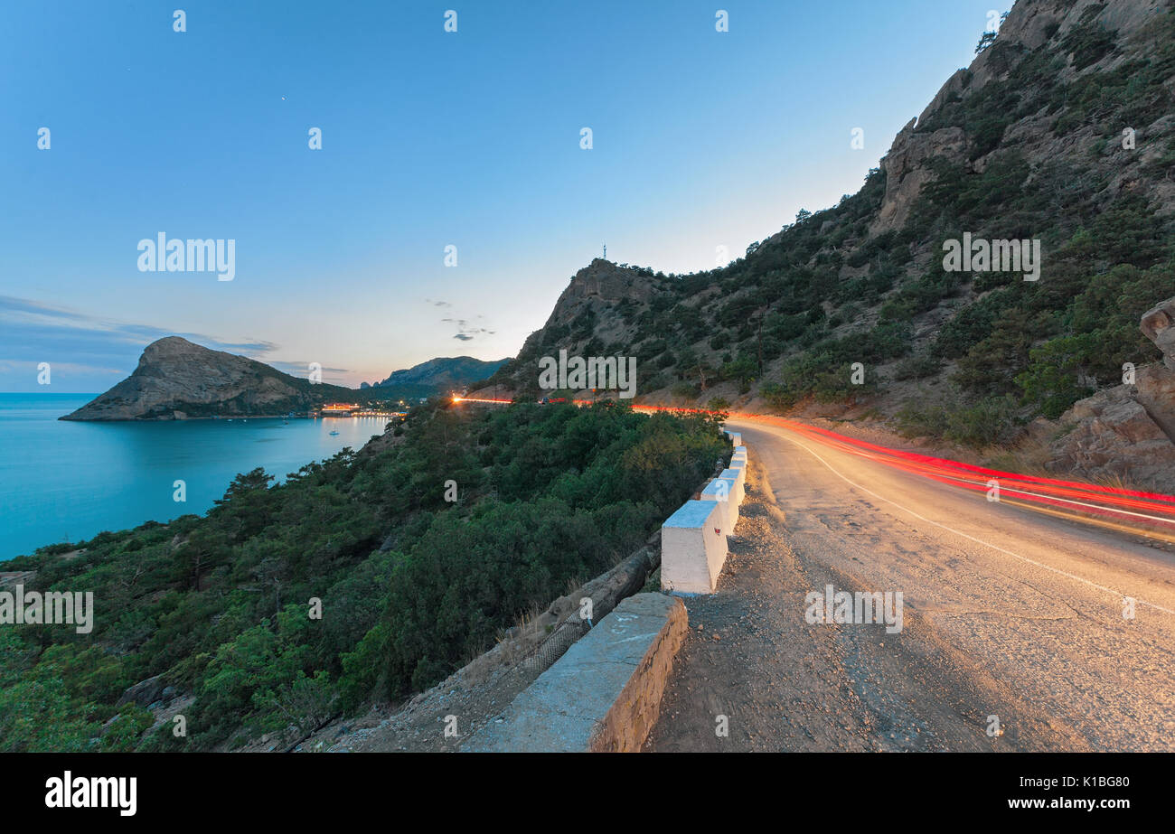 Die Serpentinen in den Bergen mit dem Licht von einem vorbeifahrenden Auto Trace in der Dämmerung auf einem Hintergrund der Berge, Strand und Meer. Stockfoto