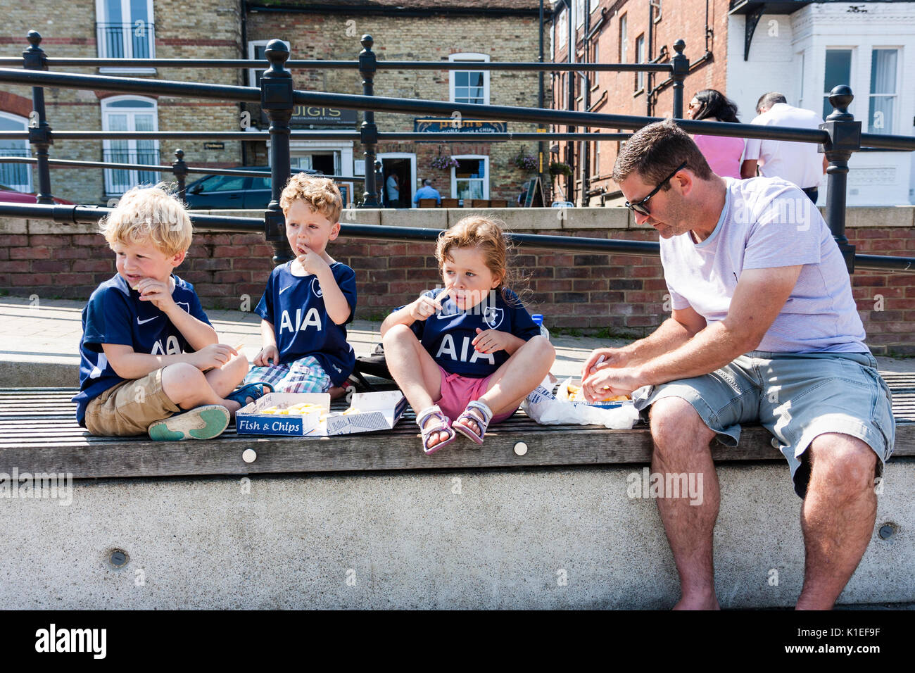 England, Sandwich. Drei junge Kinder, 5-6 Jahre alt, mit ihrem Vater, sitzen an der Wand in einem Feld von Fisch und Chips im Sommer strahlender Sonnenschein. Stockfoto