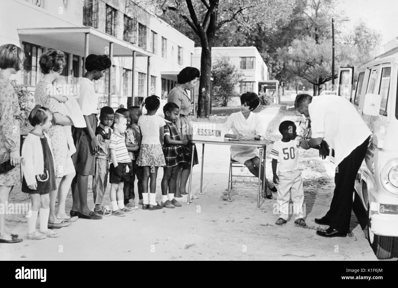 Dieses 1976 Foto zeigt eine multiethnische Gruppe Frauen und Kinder in einem Gehäuse Projekt Futter bis zu einer mobilen Klinik für das Warten und das Empfangen von Impfungen. Die Szene enthält auch einen Arzt, der die Verwaltung der Einspritzungen wurde, und eine Krankenschwester, die Registrierung der Kinder durch elterliche Beteiligung. Für eine Farbe Version dieses Bild, siehe PHIL 17503. Dieses Szenario war in einem hypothetischen Dixon Lenkstange County, USA entfernt worden und wurde als Lehrmittel im Hinblick auf die Prävention von Impfstoff - vermeidbare Krankheiten verwendet. Mit freundlicher CDC/Reguel Waldrop, 1972. Stockfoto