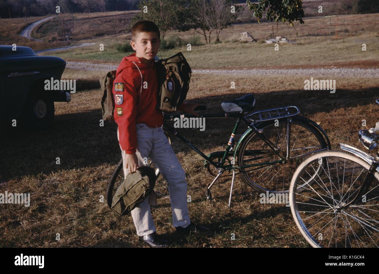 Ein in voller Länge Porträt eines Jungen tragen ein rotes Langarmshirt mit Patches, beladen mit zwei Taschen und stand neben seinem geparkten Fahrrad in eine Rasenfläche neben eine kurvenreiche Straße, 1966. Stockfoto