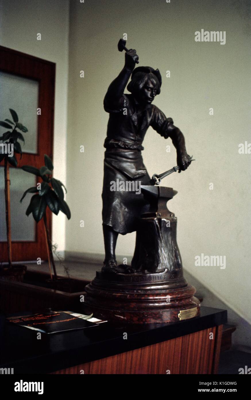 Eine kleine Statue von einem Schmied arbeiten ein Stück Metall auf einen Amboss, als Dekoration in einen Eingangsbereich mit Pflanzen im Hintergrund, 1966. Stockfoto