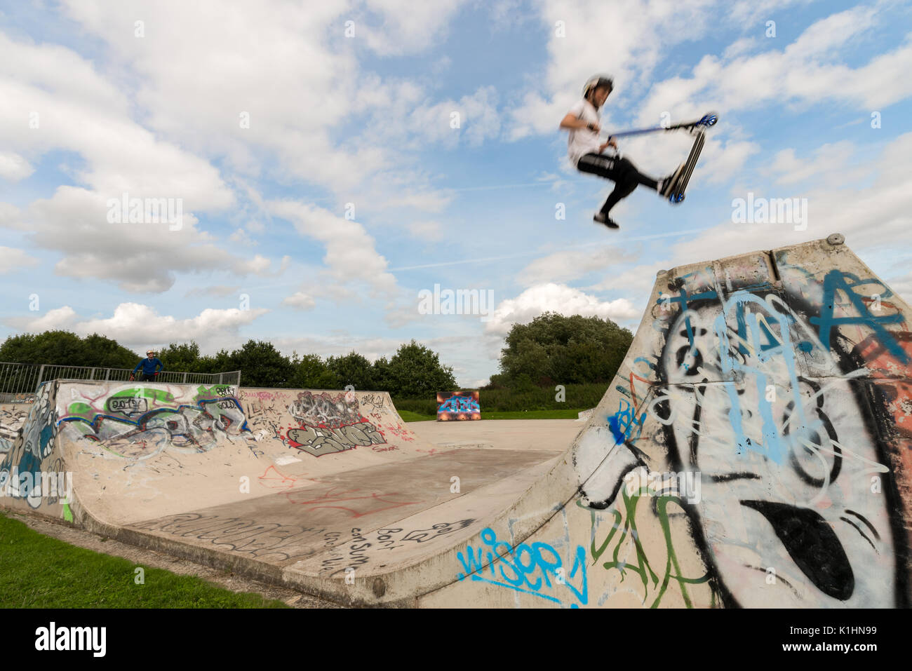 Ein Jugendlicher auf einen Roller hat einen Sprung Trick auf eine konkrete Halfpipe Rampe von Stratford-upon-Avon Skate Park während der Sommerferien. Stockfoto