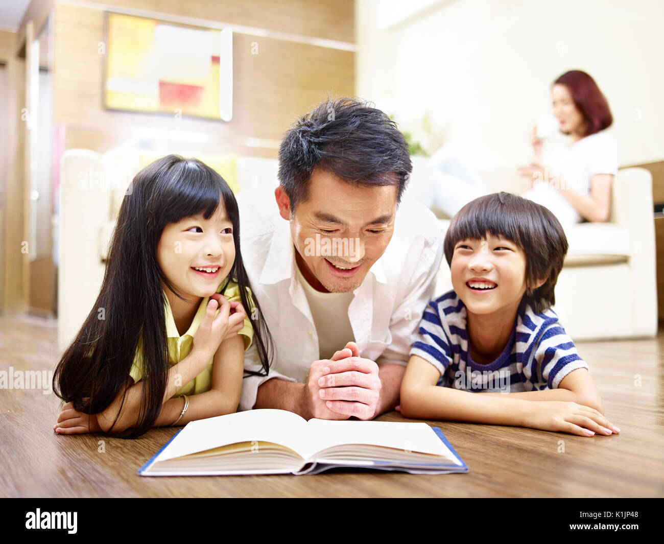 Asiatische Vater und Kinder auf dem Boden lesen Buch liegen mit Mutter im Hintergrund. Stockfoto