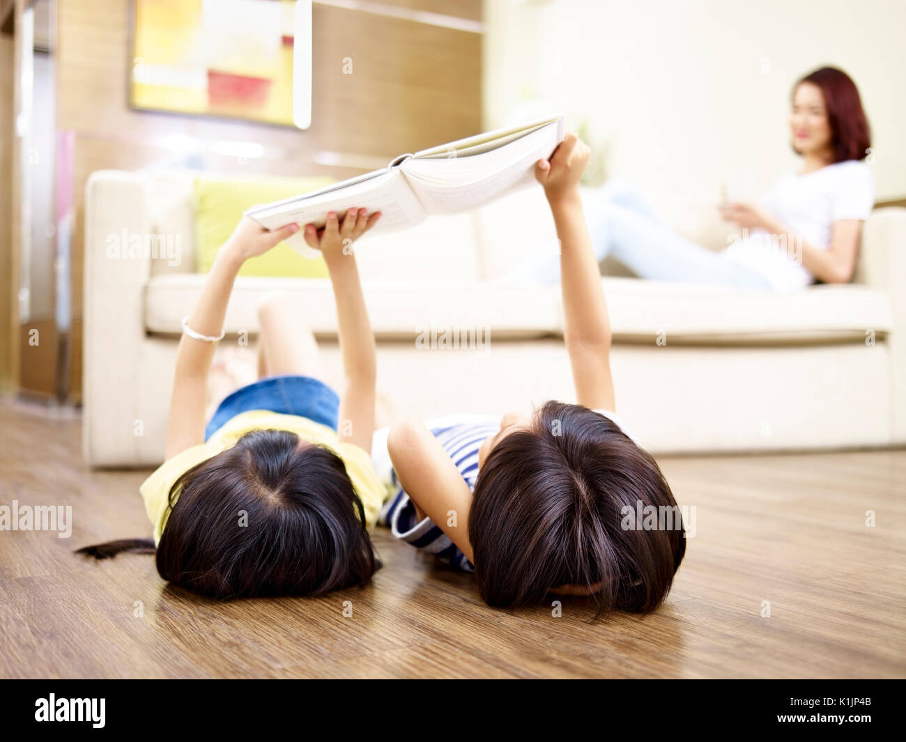 Asiatische Kinder liegen auf dem Rücken auf dem Boden mit digitalen Tablet mit Mutter sitzen auf der Couch im Hintergrund. Stockfoto