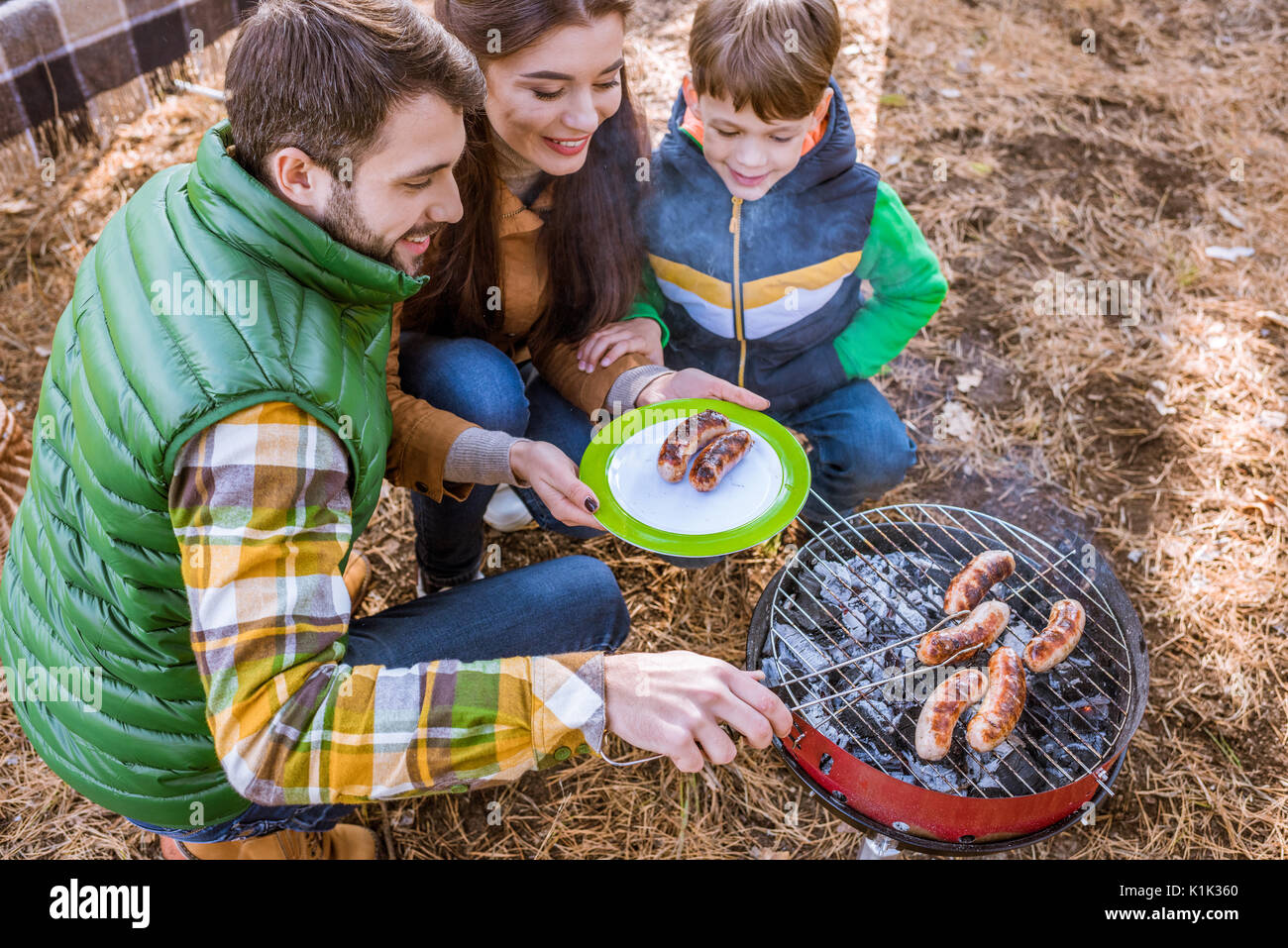 Fröhliche Eltern mit niedlichen kleinen Sohn grillen Würstchen auf Grill im Herbst Park Stockfoto