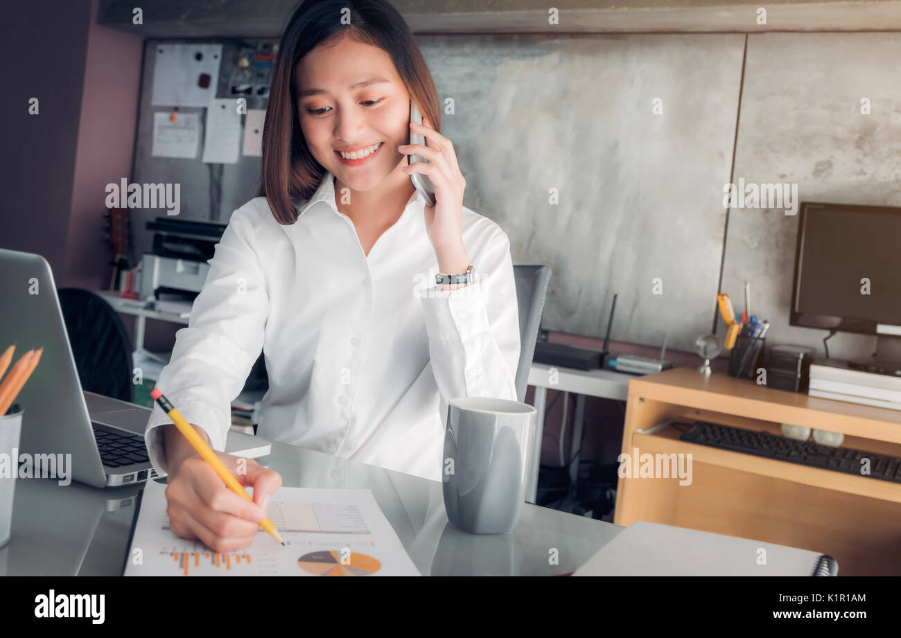 Asiatische Geschäftsfrau mit lächelnden Gesicht sprechen auf mobilen und Schreiben von Daten auf Blatt auf dem Schreibtisch mit Kollegen am Telefon analysieren, Büro Leben anhand von quantitativen Simulatio Stockfoto