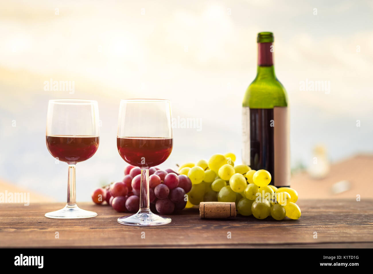 Zwei Gläser guten Rotwein mit Trauben, Flasche und Korken auf einen hölzernen Tisch während einer romantischen Sommer outdoor Diner Stockfoto