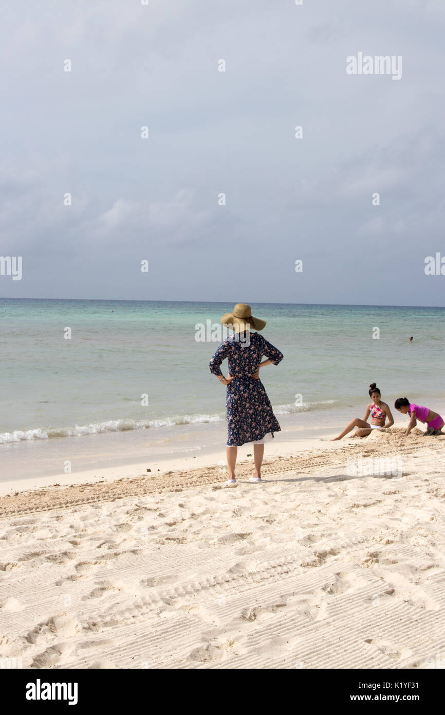 Asiatische Frau mit Schlapphut und einfachen 3/4-Ärmel Kleid auf den Hüften an stürmischen Karibik suchen. Von hinten gesehen. Stockfoto