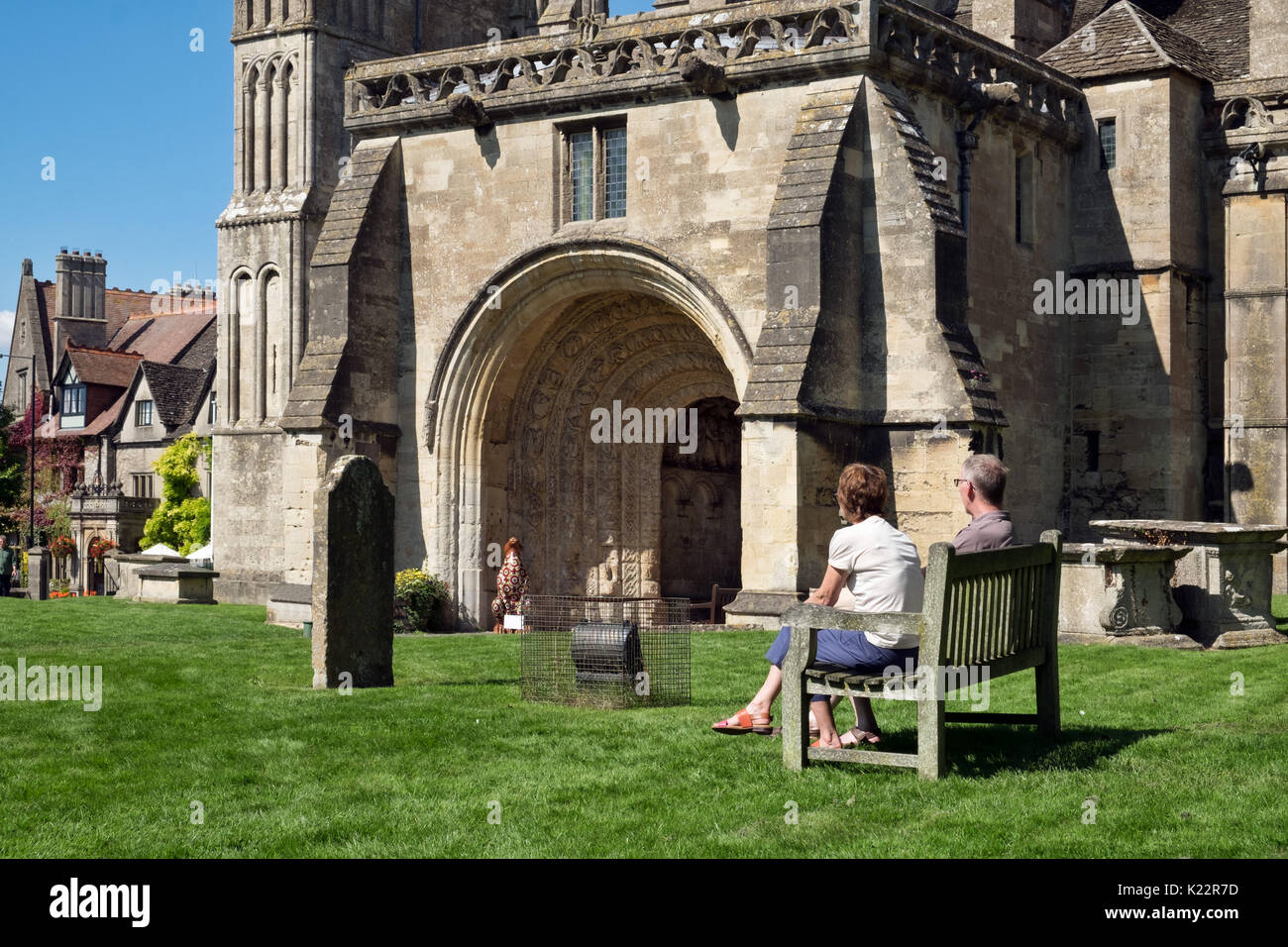 Ein paar der Besucher auf einer Bank sitzen, an einem sonnigen Tag auf dem Friedhof aus dem 7. Jahrhundert Malmesbury Abbey an einem sonnigen Tag in Wiltshire, Großbritannien Stockfoto