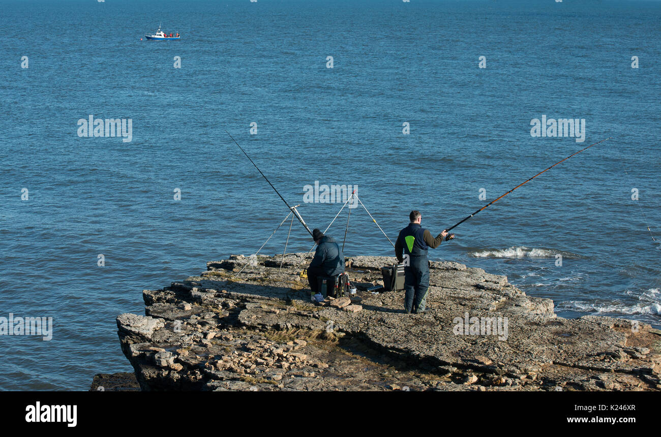 Zwei Angler mit Rod- und Linien-Seeangeln von Küstenklippen in Northumberland mit einem kleinen Boot und Seeanglern im Hintergrund Stockfoto