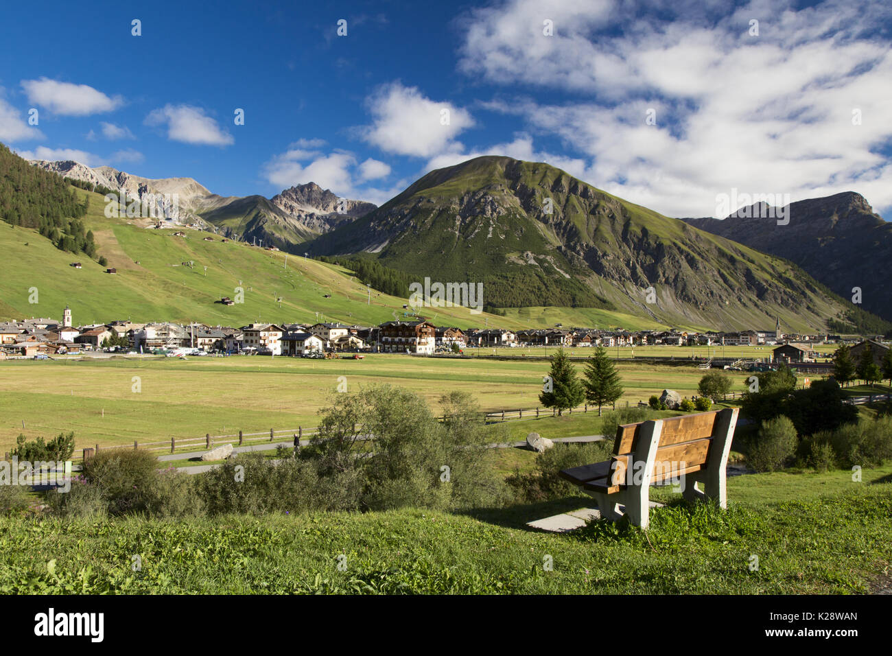 Große Stadt in der Ferne und Holzbank Nahaufnahme mit hohen grünen Bergen im Hintergrund und bewölktem Himmel Stockfoto