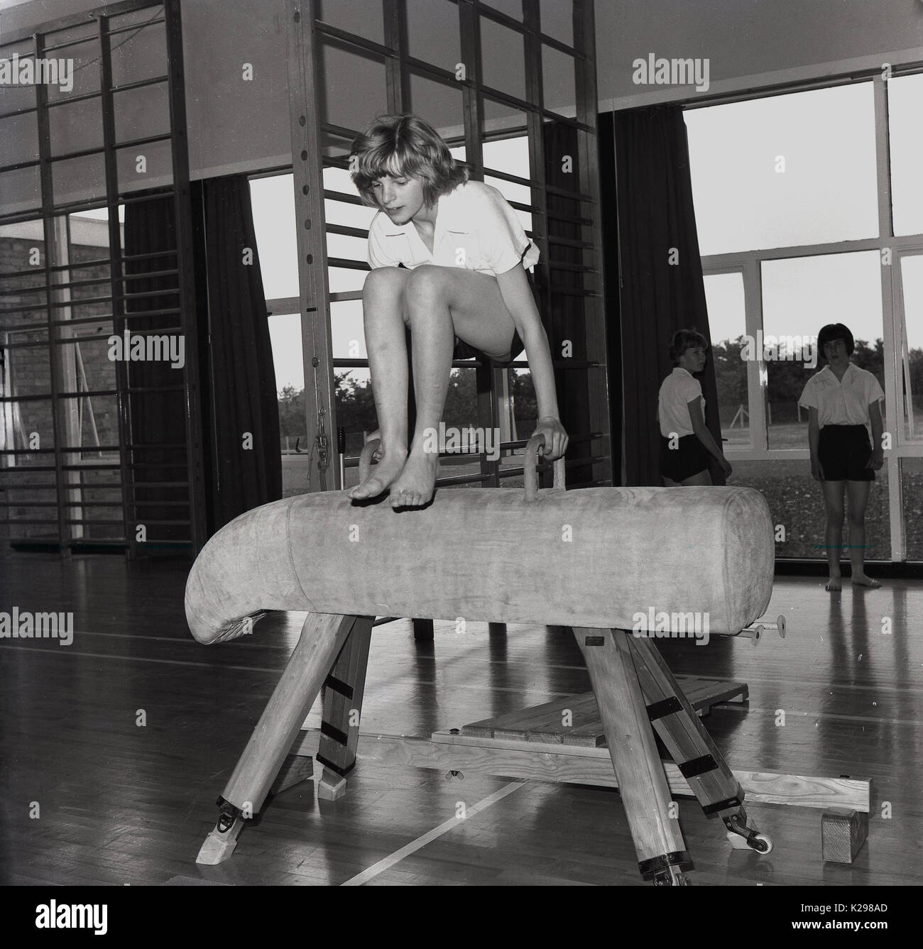 1960, historische, Bild zeigt ein junges Mädchen in einer weiterführenden Schule springen oder vaulting in Barfuss über eine Wildleder bedeckt Holz gerahmt Pferd in der Schule Hall, England, UK. Stockfoto
