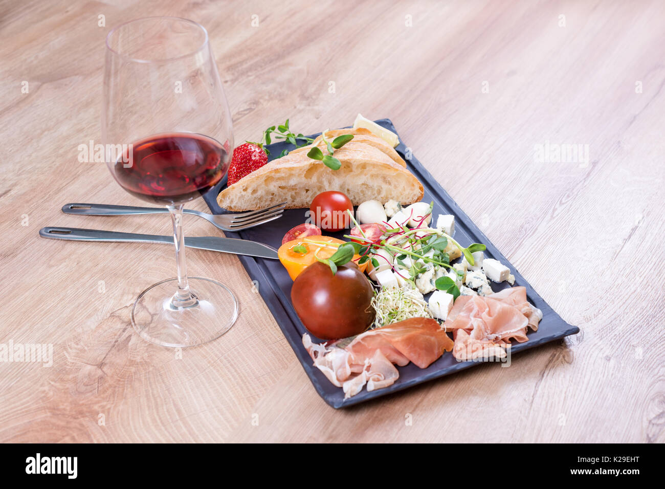 Snacks mit Rotwein auf schwarzem Schiefer an Bord über hölzerne Hintergrund. Delikatesse und einfaches Essen, Brot, Käse, Tomaten und Schinken. Stockfoto