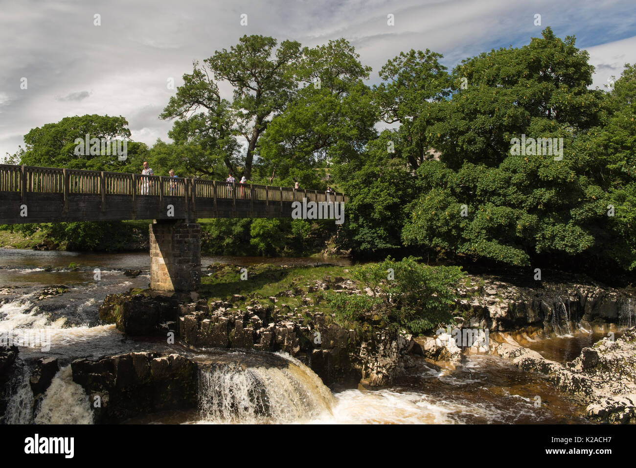 Sonnigen Sommertag & Menschen kreuzen Holzsteg über malerische Linton fällt Wasserfall - River Wharfe, Grassington, Yorkshire Dales, England, UK. Stockfoto