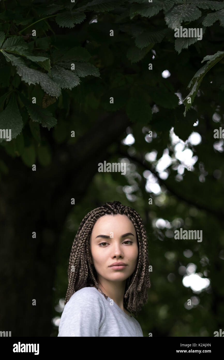 Ein schönes Mädchen mit Afro Pigtails blickt ernst in die Kamera auf einem Hintergrund von grünem Laub der Bäume. Stockfoto