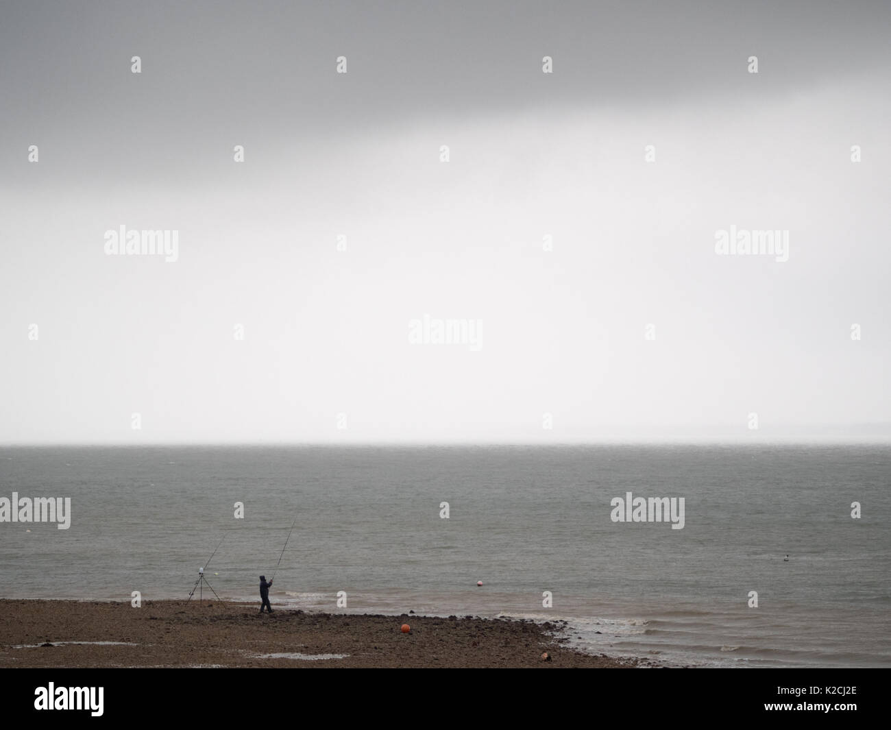 Ein einsamer Fischer Silhouette der Fischerei durch die Kante des Solent sea in Bad regnet Wetter mit Stangen von Strand unter schweren Grau Grau dark sky Stockfoto