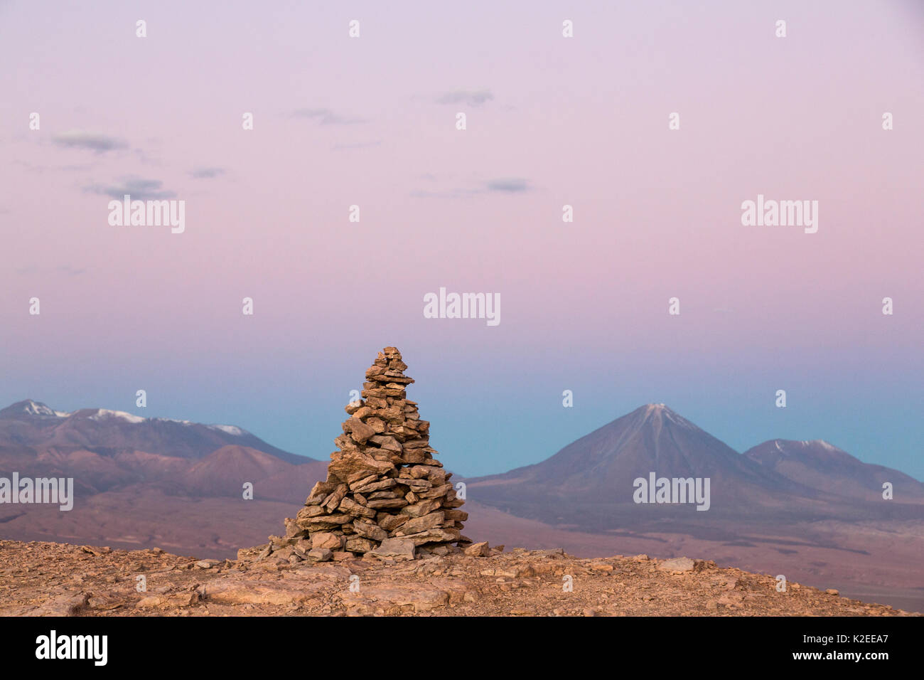 Apacheta/Apachita - ein Stein Cairn in den Anden, ein kleiner Haufen von Steinen auf dem Weg in den hohen Bergen mit Licancabur Vulkan gebaut (5916 m über dem Meeresspiegel) und Juriques Vulkan auf der rechten Seite (5704 m) im Hintergrund in der Dämmerung, Atacama, San Pedro, Chile Stockfoto