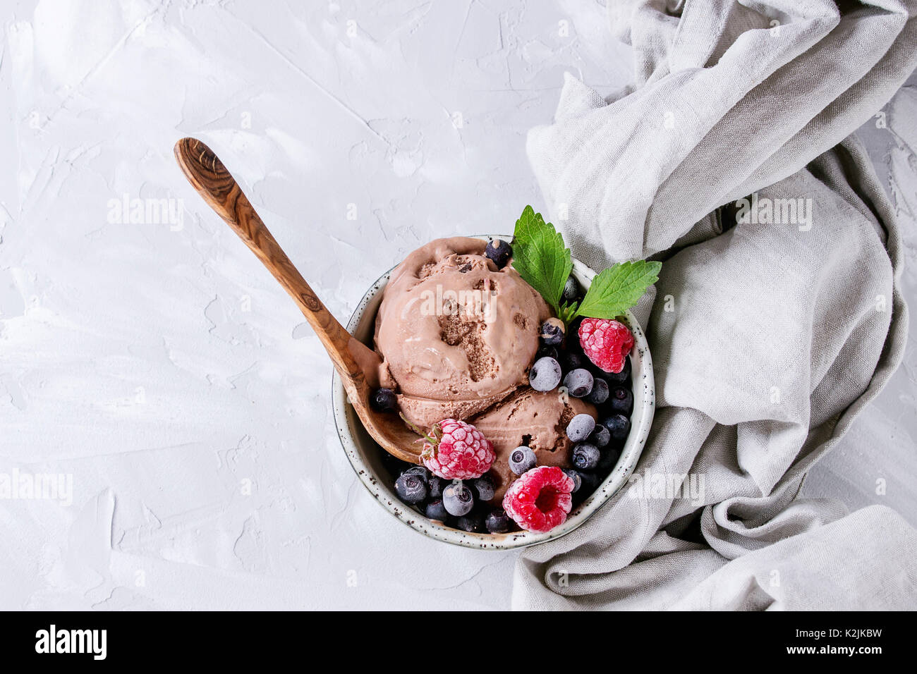 Hausgemachten Schokoladeneis mit gefrorenen Beeren Heidelbeere, Himbeere, Minze, serviert in weiße Schüssel mit Olivenholz Löffel und textilen Leinen über Graues con Stockfoto