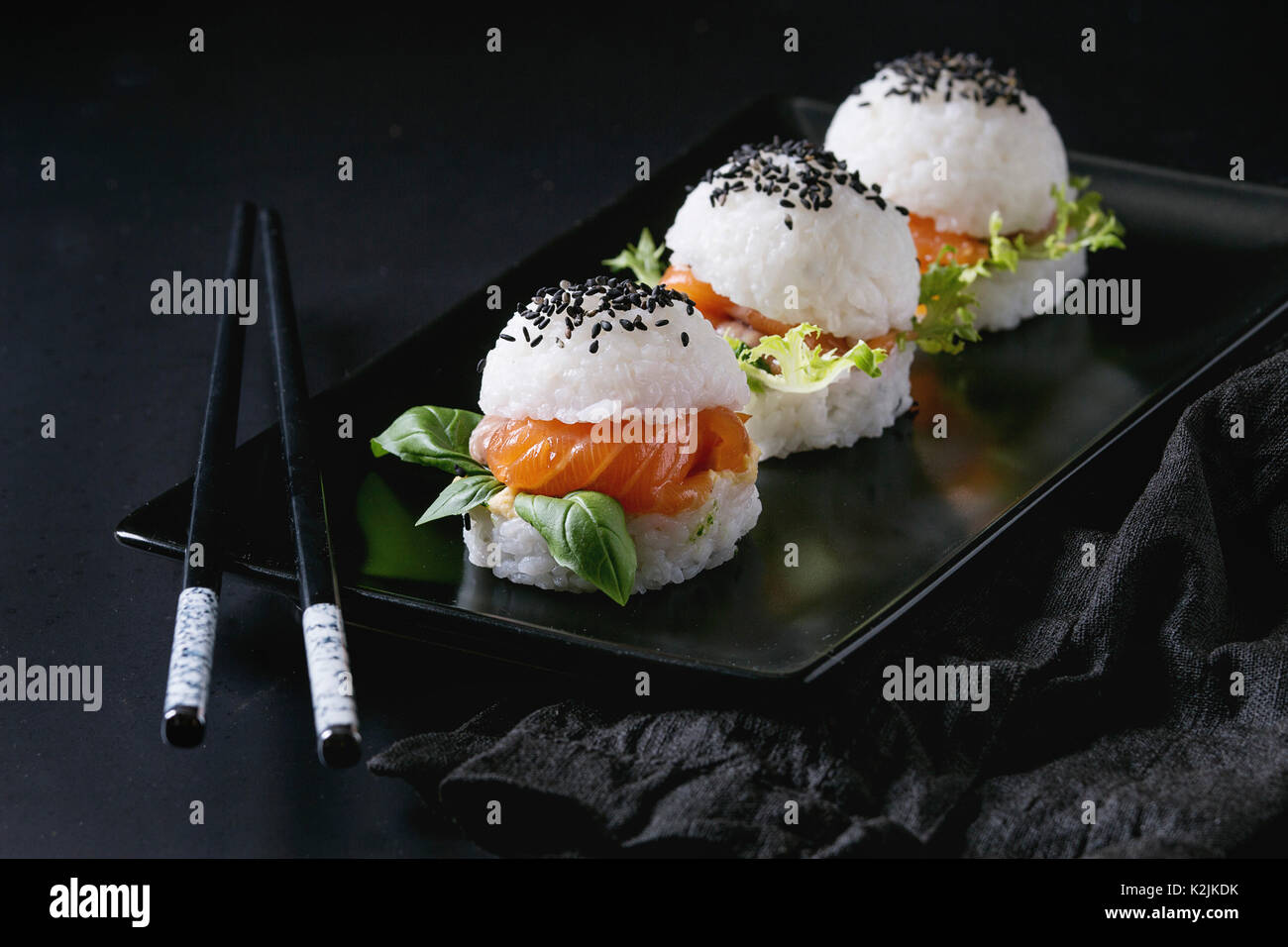 Mini Reis sushi Burger mit geräuchertem Lachs, grüner Salat und Saucen, schwarzer Sesam auf Schwarze quadratische Platte mit Stäbchen und textile Serviette serviert über Stockfoto