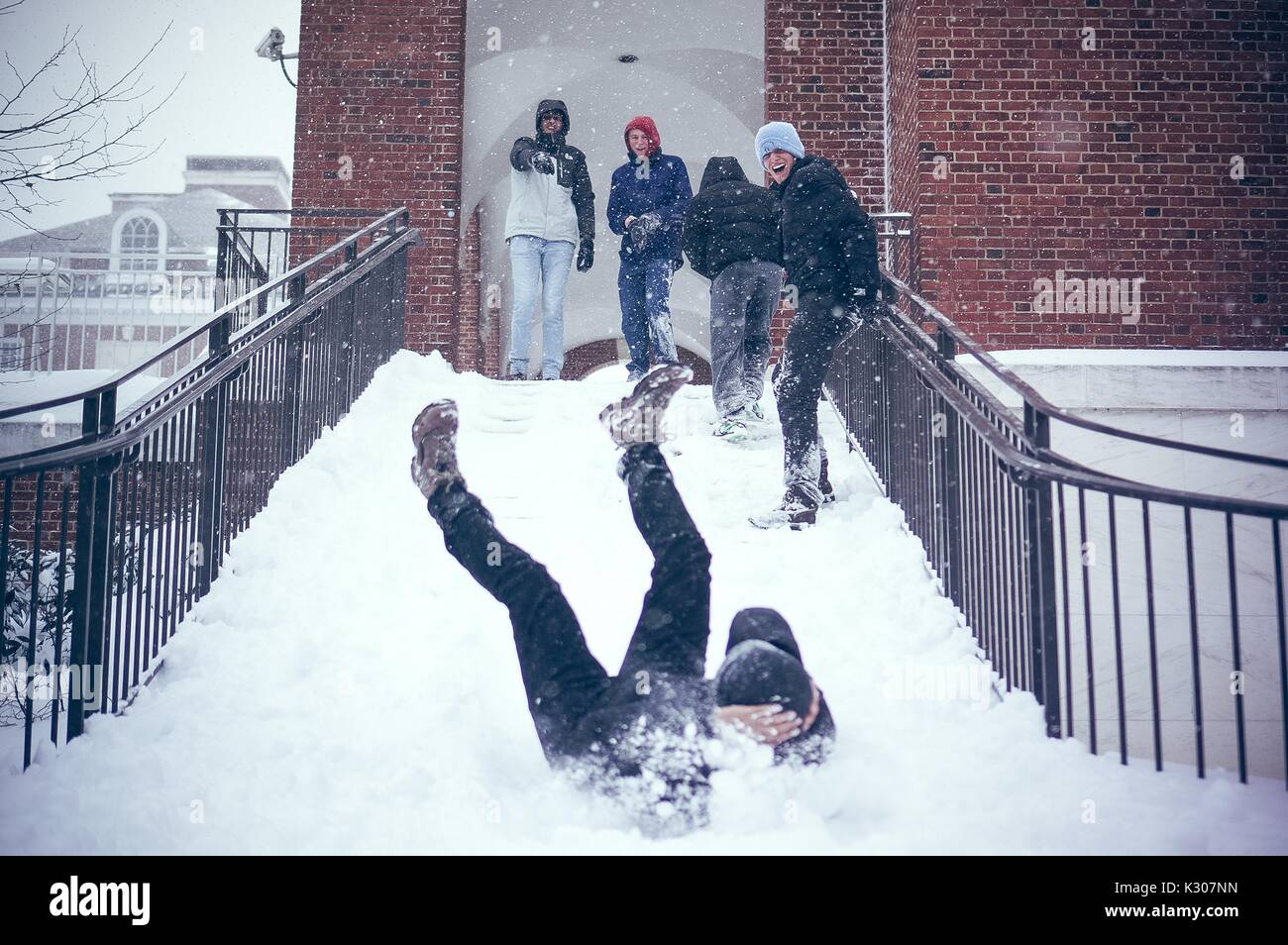 Ein Student rutscht einem verschneiten Treppen mit Beine in die Luft und Arme über seinen Kopf, während andere Studenten im Schnee gang Lächeln und Jubeln am oberen Ende der Treppe, auf einem Snow Day an der Johns Hopkins University, Baltimore, Maryland, 2016. Stockfoto