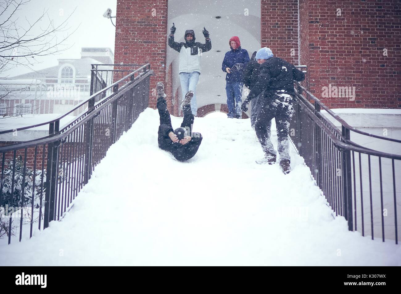 Ein Student Rutschen rückwärts mit Beinen in der Luft und die Arme um seinen Kopf, unten eine verschneite Treppe, während Freunde im Schnee gang Beifall von oben an der Treppe, während einer Snow Day an der Johns Hopkins University, Baltimore, Maryland, 2016. Stockfoto