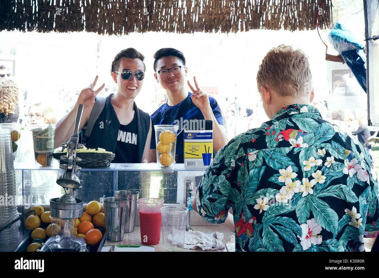 Zwei Studenten pose mit peace Zeichen hinter dem Tresen einer tiki-Stil Limonade Stand, während ein Mann in Hawaiian shirt ihre Getränke vorbereitet, während der Spring Fair, ein Kursteilnehmer - Spring Carnival an der Johns Hopkins University, Baltimore, Maryland, April, 2016. Mit freundlicher Genehmigung von Eric Chen. Stockfoto