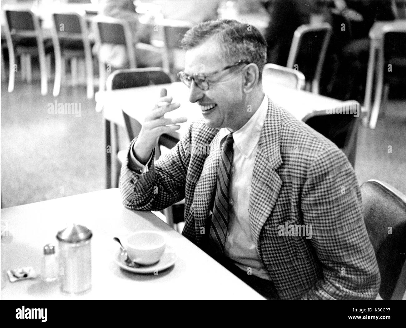 Tragen tweed Blazer und Tortoise Gläser, Thomas Ira Koch, Professor für Politische Philosophie an der Johns Hopkins University, sitzt an einem Tisch in einem Diner seine Hand halten bis zu seinem Gesicht beim Lachen, eine leere Kaffeetasse vor ihm, Baltimore, Maryland, 1956. Stockfoto