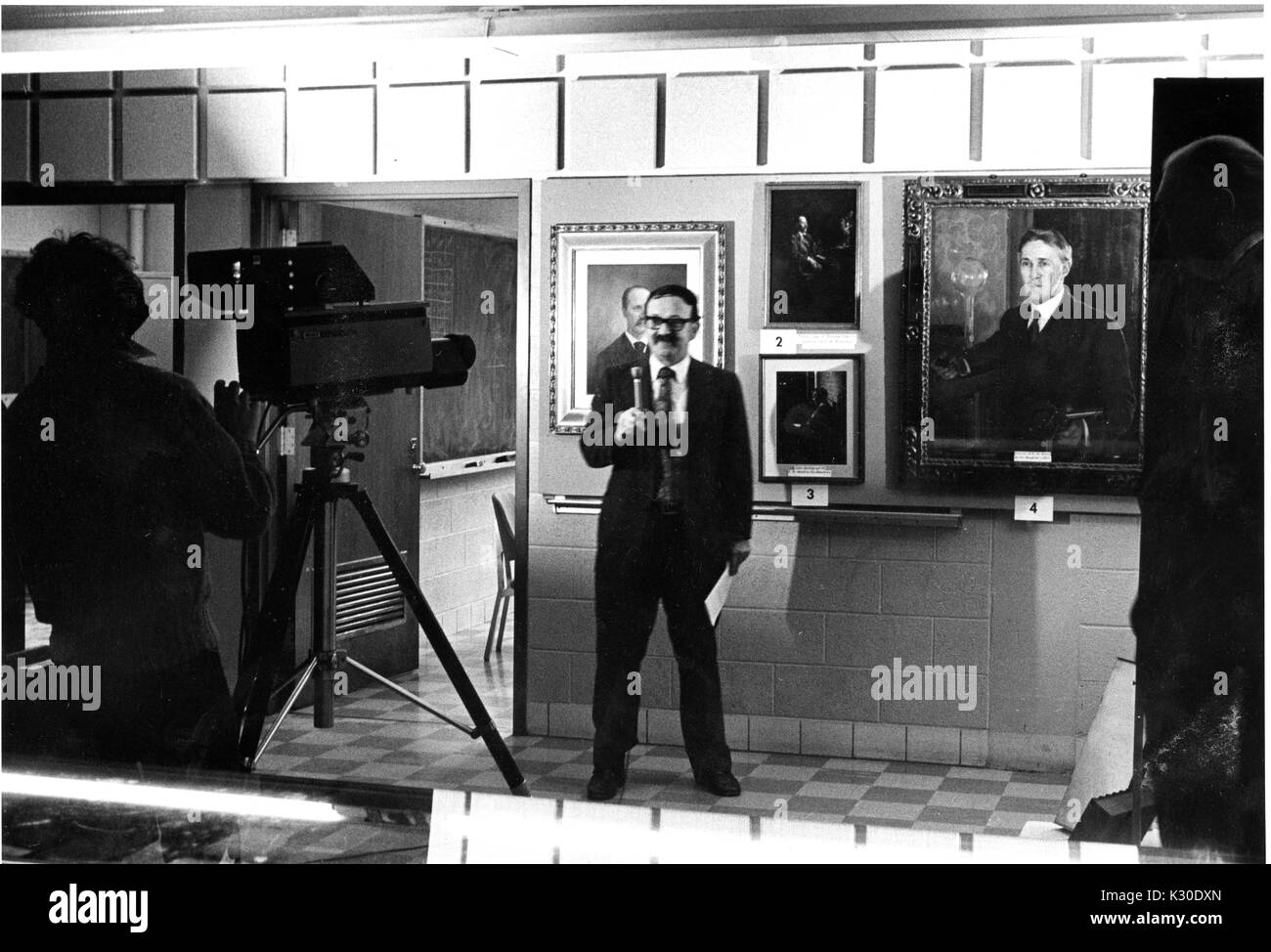 Ein Mann mit einem Mikrofon und einem Schnurrbart und Gläser vor Porträts von Henry Rowland und Robert Holz bei Rowland-Wood Symposium, 1976, gefilmt zu werden. Stockfoto