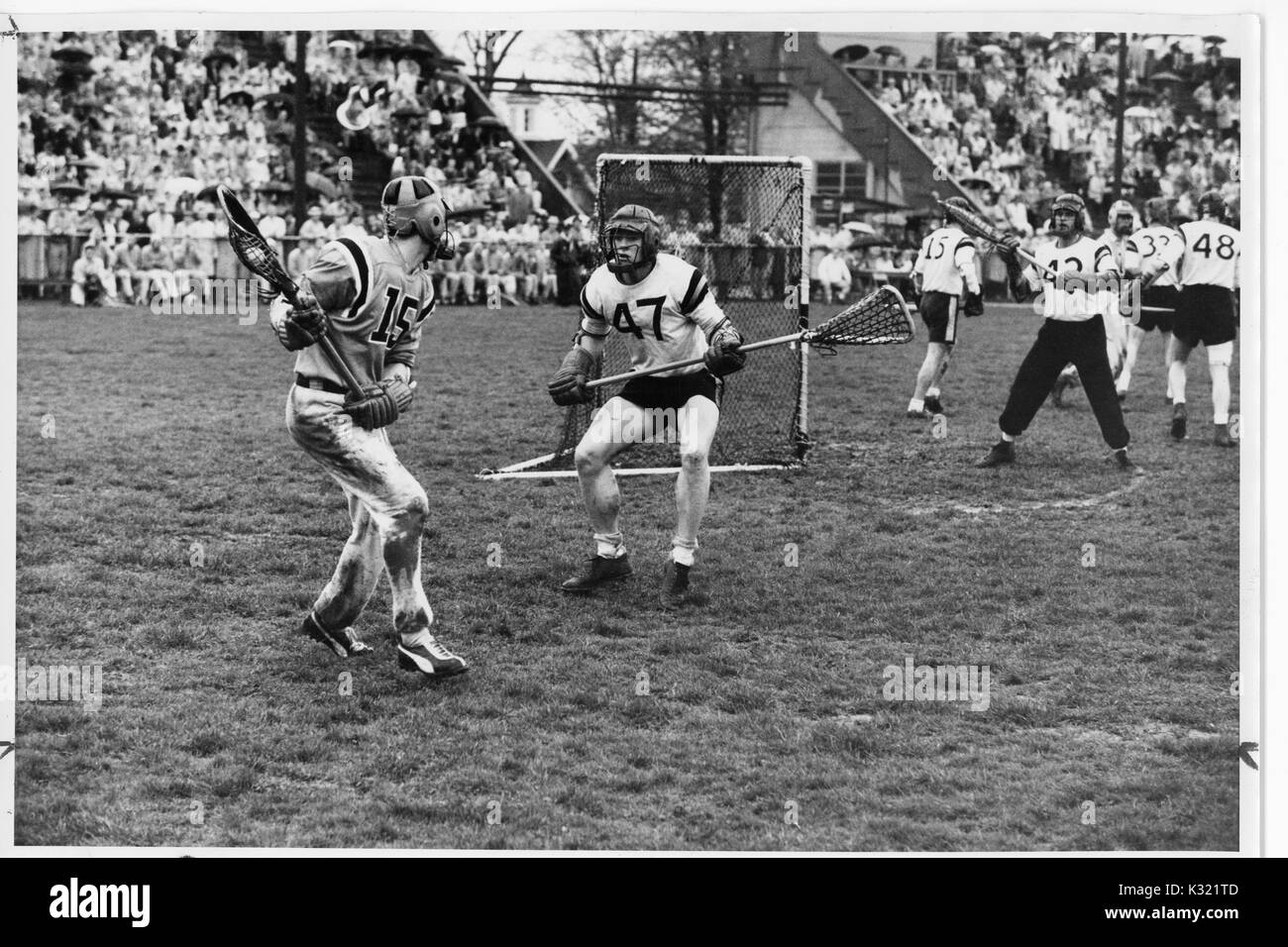Graustufen Aktion Foto während einen Men's lacrosse Übereinstimmung zwischen der Johns Hopkins University und der Princeton University, als JHU Player bereitet einen Pass von hinter dem Tor, während die gegnerische Spieler versuchen, seine Bewegung zu blockieren, so überfüllt steht voller Leute beobachten, die das Spiel, Baltimore, Maryland, Juni, 1959. Stockfoto