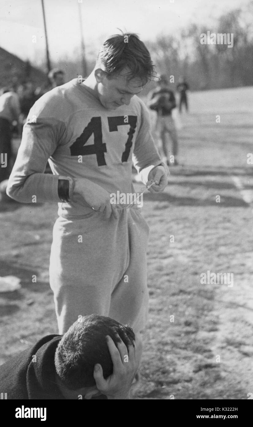 Sepia Ton offen Foto von Byron Forbush Nummer 47, Co - Kapitän von der Men's lacrosse Mannschaft an der Johns Hopkins University, einheitliche und ständigen Tragen am Rand eines Feldes einstellen seine Trainingshose Tunnelzug, während ein anderes Teammitglied unten kauert auf dem Feld vor ihm, Baltimore, Maryland, Juni, 1951. Stockfoto