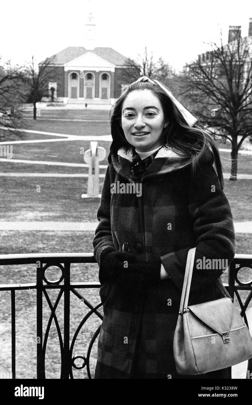 Eine weibliche Schulabgänger an der Johns Hopkins University, das Tragen von Wintermantel und Geldbeutel von ihrem Handgelenk, lächeln und stellt für Ihr Foto auf der Wandelgang mit Blick auf Wyman Quad, Baltimore, Maryland, 1975. Stockfoto