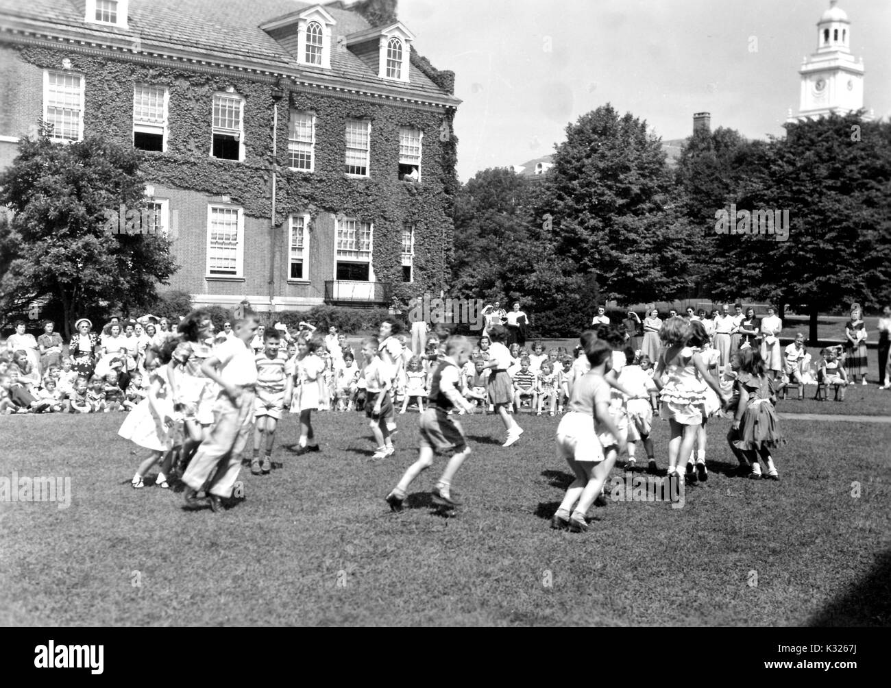 Am Ende des Schuljahres für eine Demonstration der Schule an der Johns Hopkins Universität, Jungen und Mädchen auf eine Show im Gras an einem sonnigen Tag, gerne überspringen in einem Kreis vor einem Publikum aus Mitschüler, Lehrer und Eltern sitzen und draußen Einer mit Efeu bewachsene Campus Gebäude, Baltimore, Maryland, Juli, 1950. Stockfoto