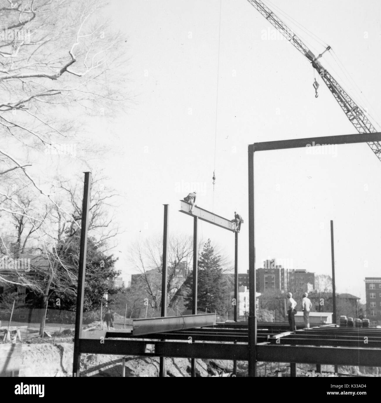 In den frühen Phasen der Konstruktion der Milton S Eisenhower Bibliothek an der Johns Hopkins University, Balken, werden eine Struktur über dem Boden zu schaffen, mit einem hohen Kran senken einen Strahl mit zwei Arbeiter sitzen auf beiden Enden, hoch in die Luft wie eine Wippe, während zwei Arbeiter Tragen von Schutzhelmen von unten anschauen, mit Bäumen und Campus Gebäude angrenzend an das Projekt Boden, Baltimore, Maryland, 1963. Stockfoto