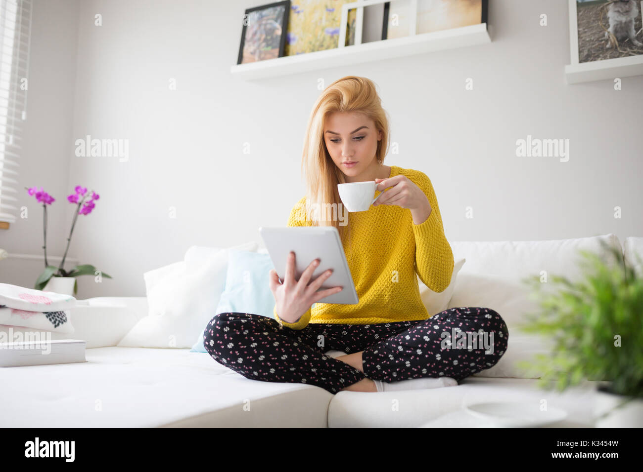 Ein Foto der jungen Frau im Schlafanzug, lesen Sie etwas Interessantes auf dem Tablett und Tee trinken. Stockfoto