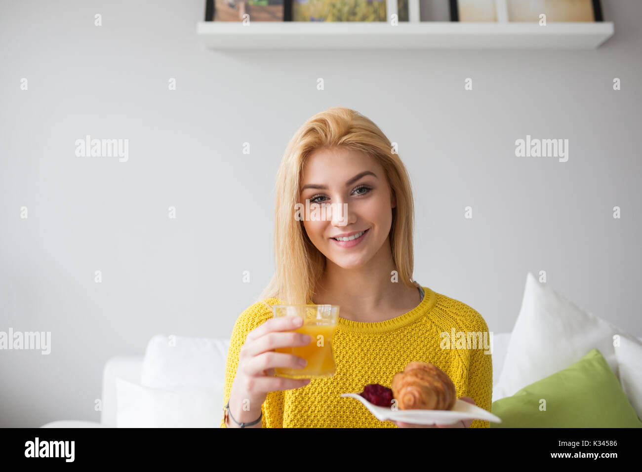 Ein Foto der jungen Frau im gelben Pullover Holding eine Platte mit Croissant und Marmelade und ein Glas Saft. Sie lächelt. Stockfoto