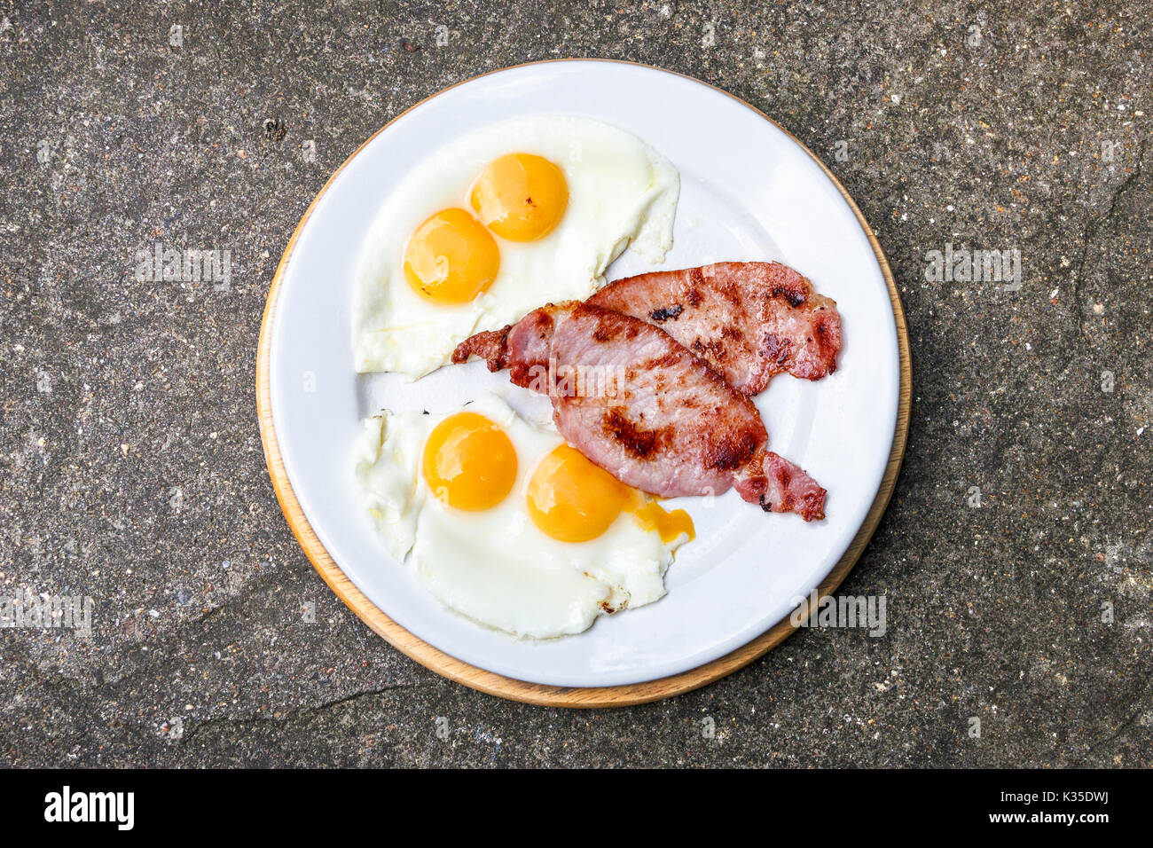 Ein traditionelles englisches Frühstück mit zwei Eier (Eigelb) mit Doppelbett und Speck auf einer weißen Platte gegen einen Stein oder Granit Hintergrund Stockfoto