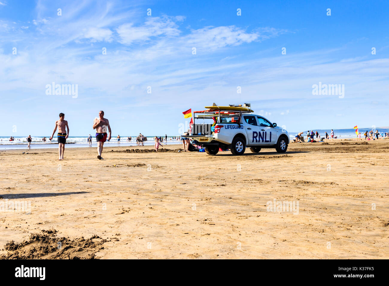 Ein rnli Rettungsschwimmer Fahrzeug und zwei männliche Urlauber Rückkehr aus dem Meer am Sandstrand bei Ebbe, Westward Ho!, Devon, England, Großbritannien Stockfoto