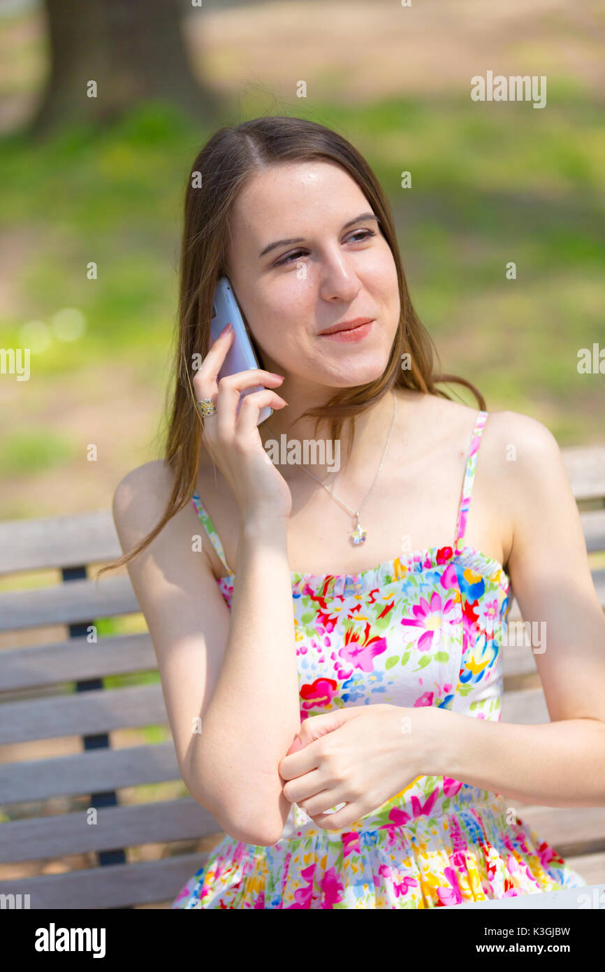 Porträt der jungen schönen Frau mit langem Haar im Sommer Park, Mädchen mit Blume kurzes Kleid mit einem -Smartphone beim Sitzen auf einer Bank Stockfoto