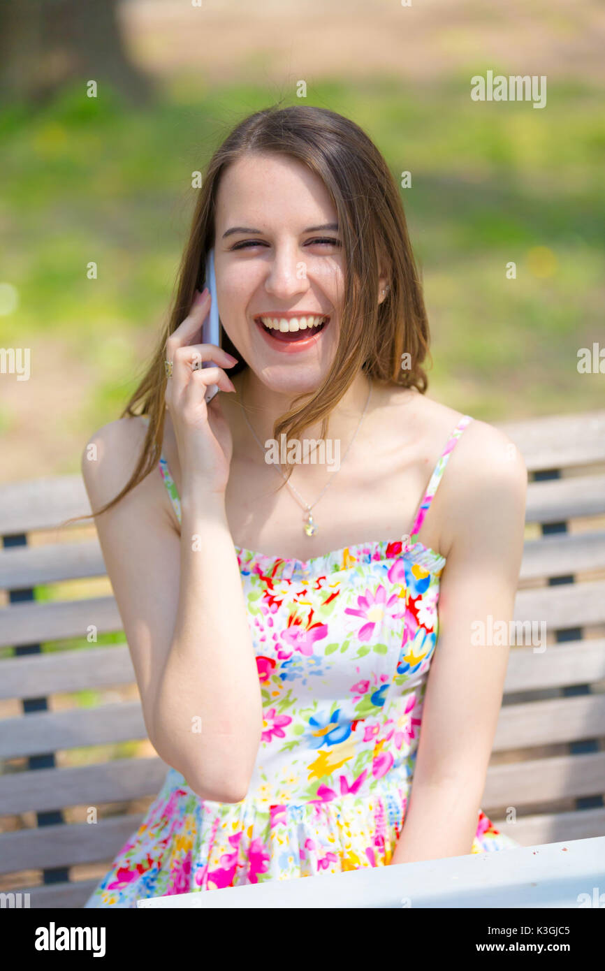 Porträt der jungen schönen Frau mit langem Haar im Sommer Park, Mädchen mit Blume kurzes Kleid mit einem -Smartphone beim Sitzen auf einer Bank Stockfoto