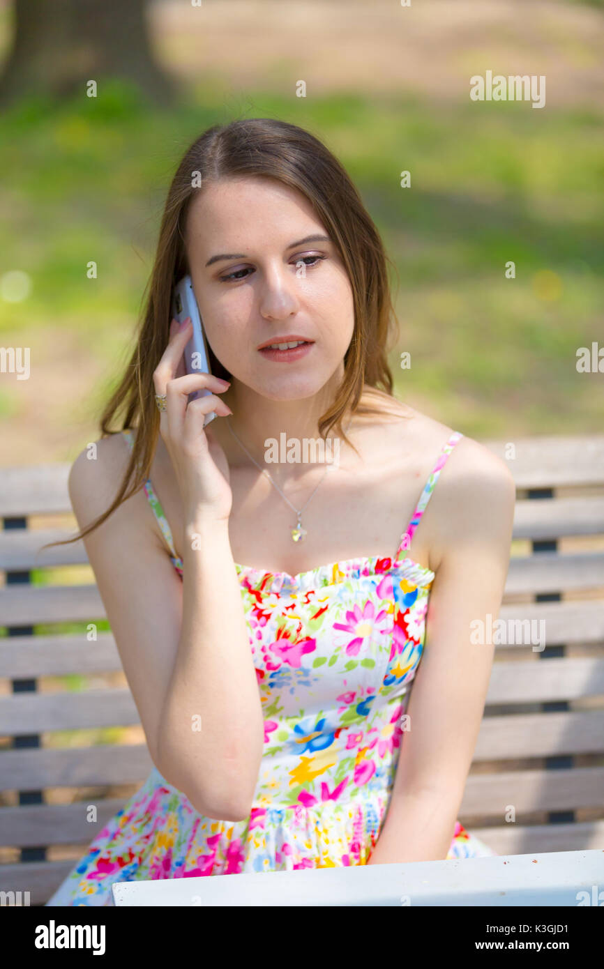 Porträt der jungen schönen Frau mit langem Haar im Sommer Park, Mädchen mit Blume kurzes Kleid mit einem -Smartphone beim Sitzen auf einer Bank Stockfoto