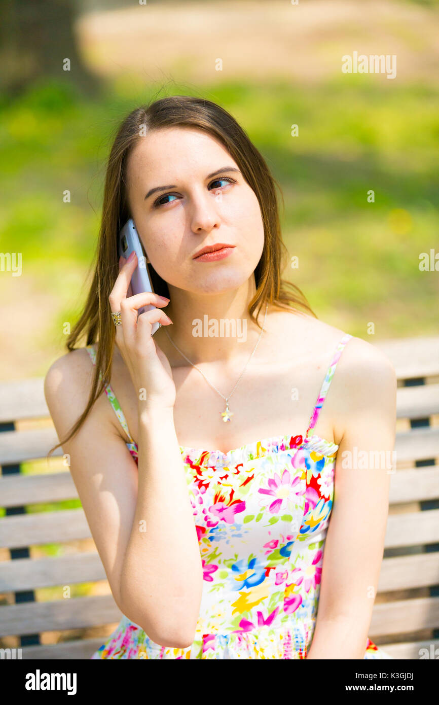 Porträt der jungen schönen Frau mit langem Haar im Sommer Park, Mädchen mit Blume kurzes Kleid mit einem -Smartphone beim Sitzen auf einer Bank Stockfoto