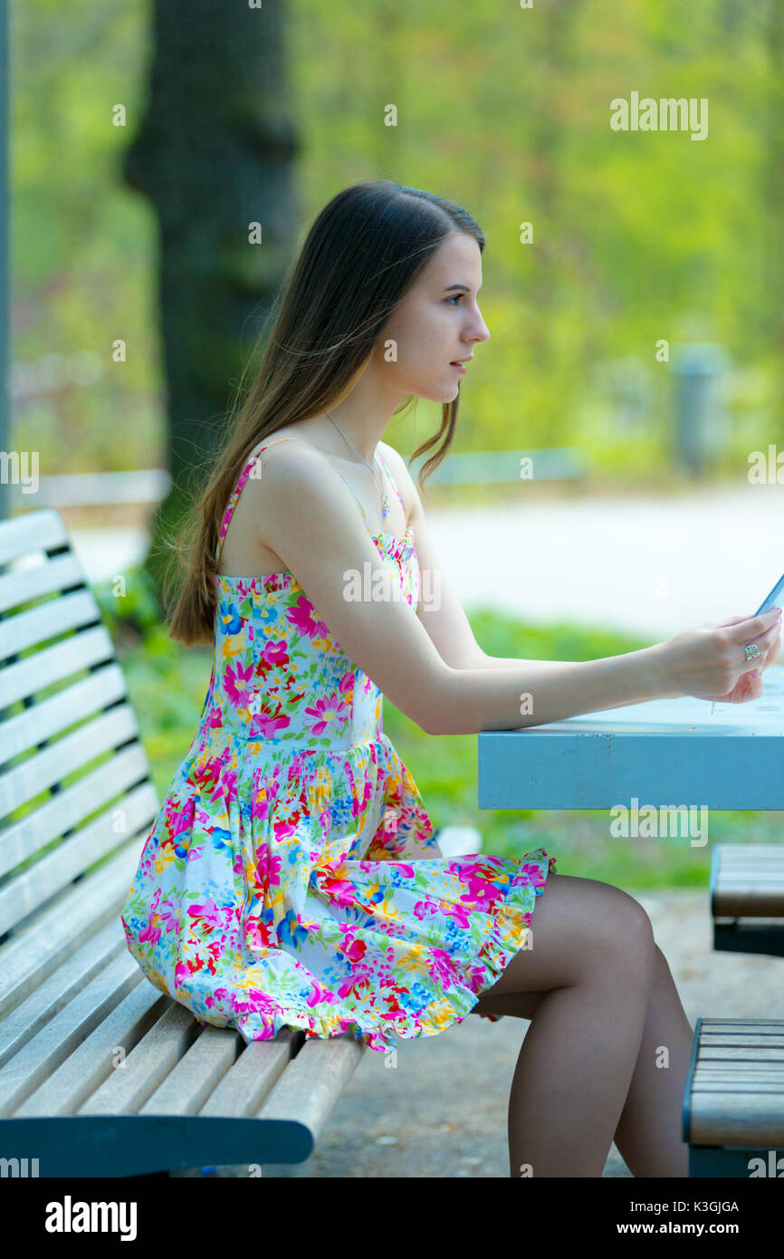 Porträt der jungen schönen Frau mit langem Haar im Sommer Park, Mädchen mit Blume kurzes Kleid mit einem -Smartphone beim Sitzen auf einer Bank Stockfoto